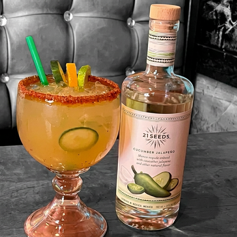 Spicy orange cocktail with cucumber garnish and chili-salt rim beside a 21 Seeds Cucumber Jalapeño tequila bottle on a restaurant table.